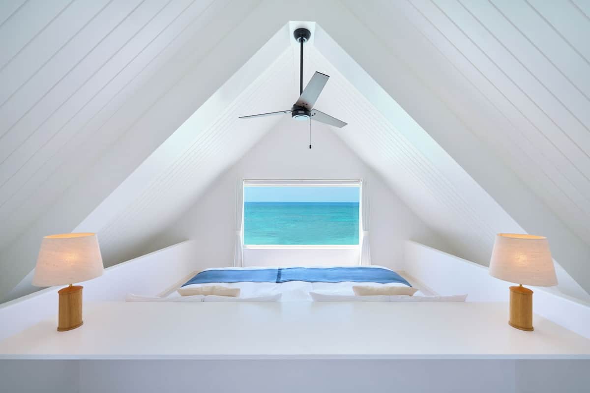 A bedroom with a sloped ceiling, a ceiling fan, a window with a view of the sea, and two bedside lamps.