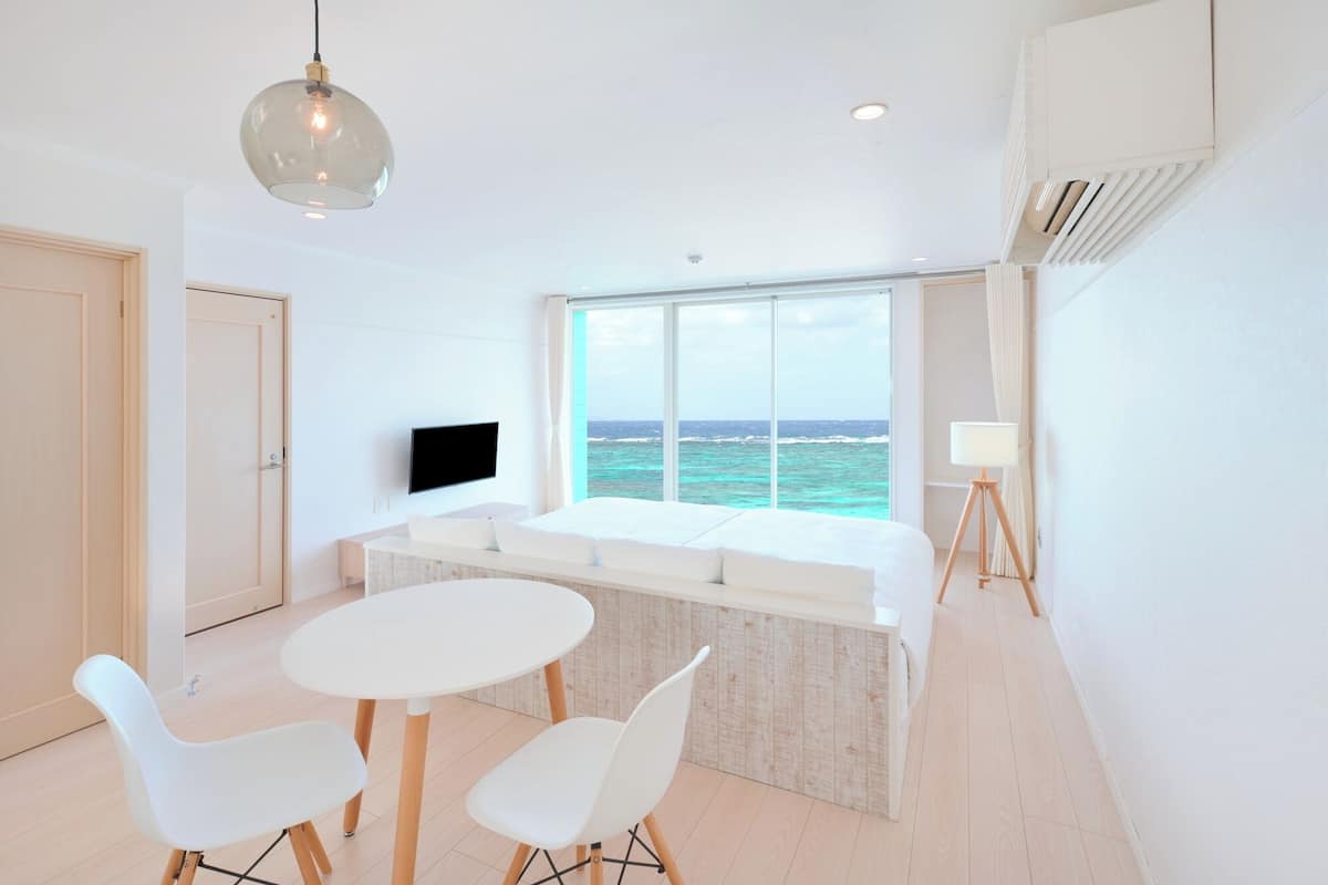 A modern room with a large window overlooking the sea, a flat-screen TV, a dining area with two chairs, and a ceiling light.