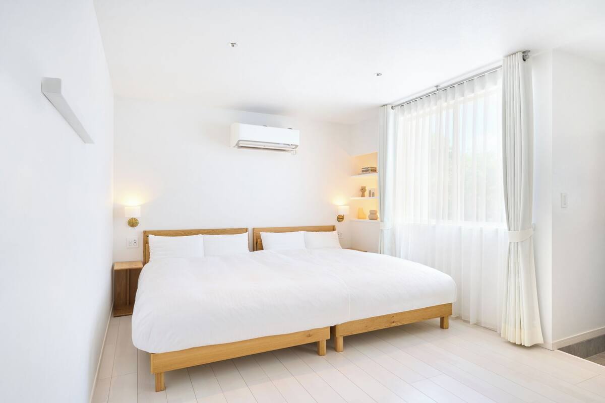 A spacious bedroom with a large bed, a bedside table, an air conditioning unit, and a window with sheer curtains.