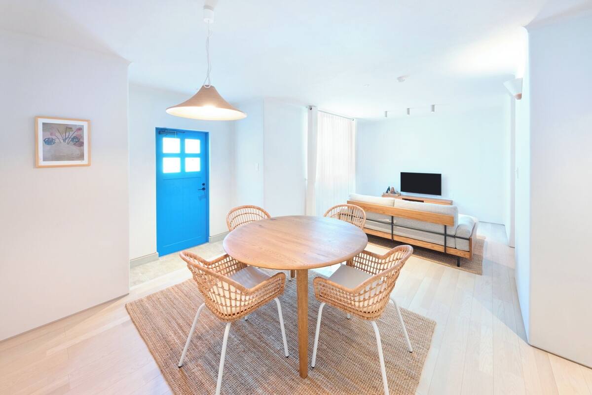 A modern living room with a round wooden table surrounded by wicker chairs, a sofa, and a flat-screen TV.