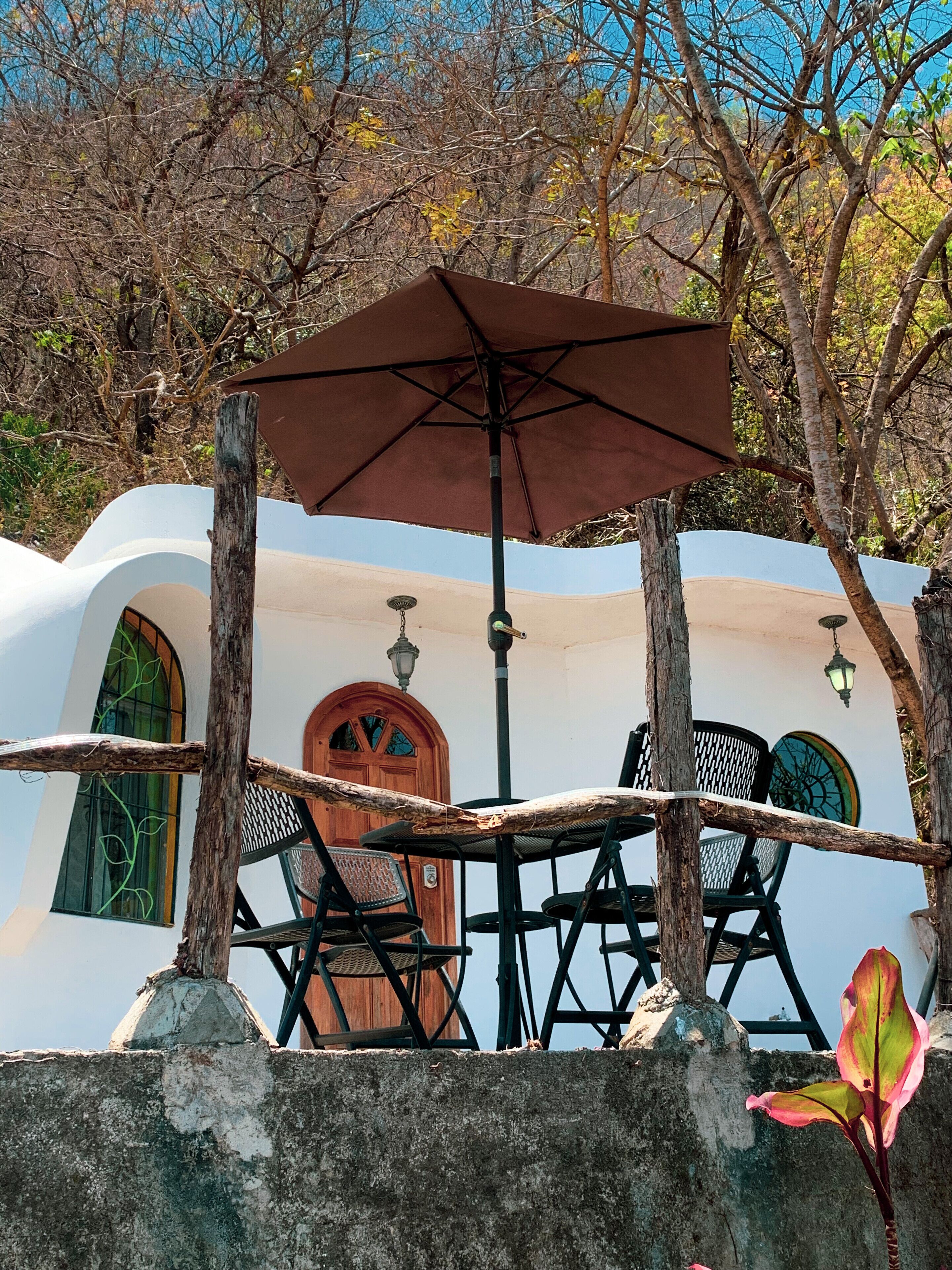 A rustic outdoor seating area with a wooden table and chairs, an umbrella, and a wall with a door and windows.