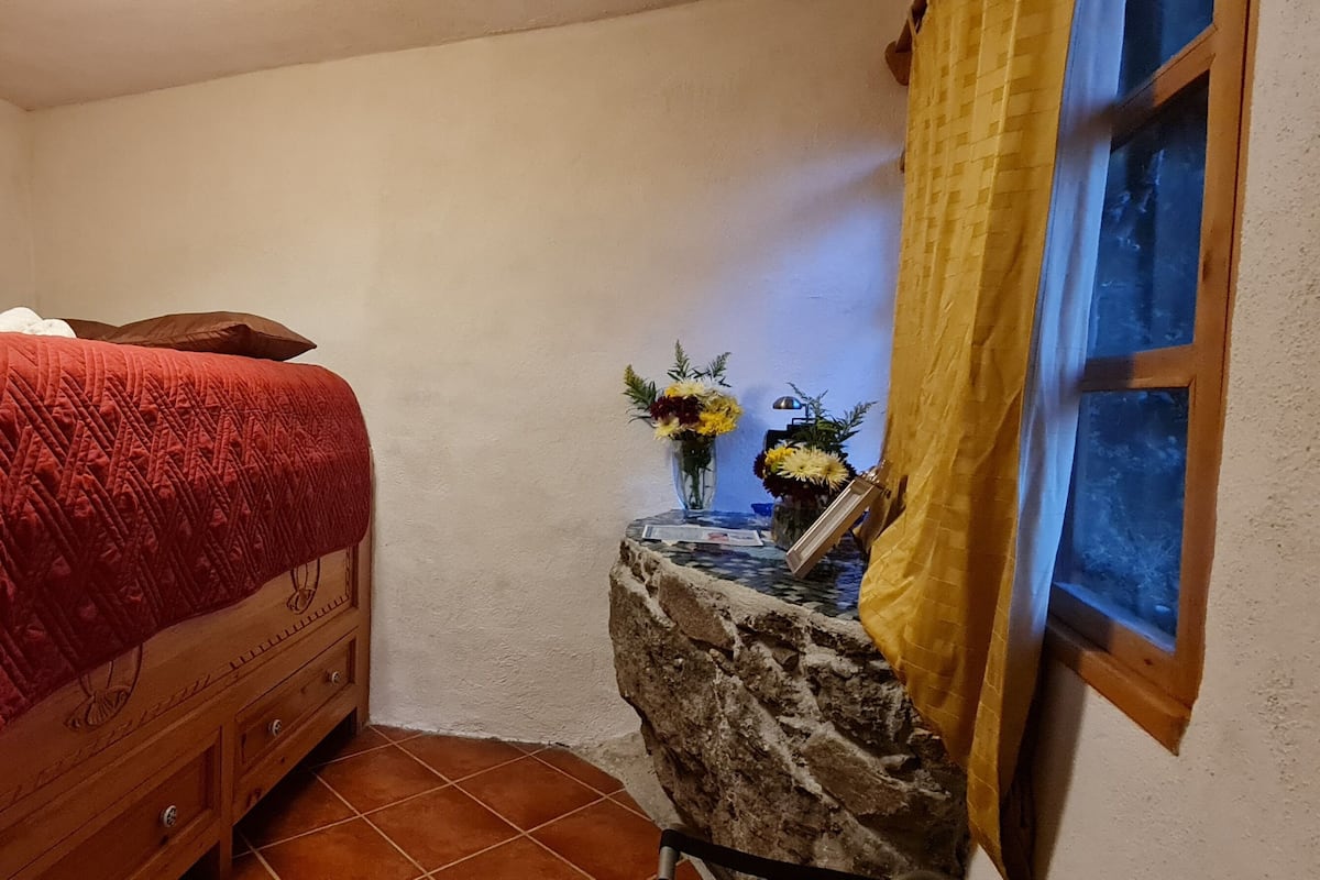 Ein Schlafzimmer mit einem hölzernen Bett, einem Nachttisch mit Blumen und einem Fenster mit gelben Vorhängen.
