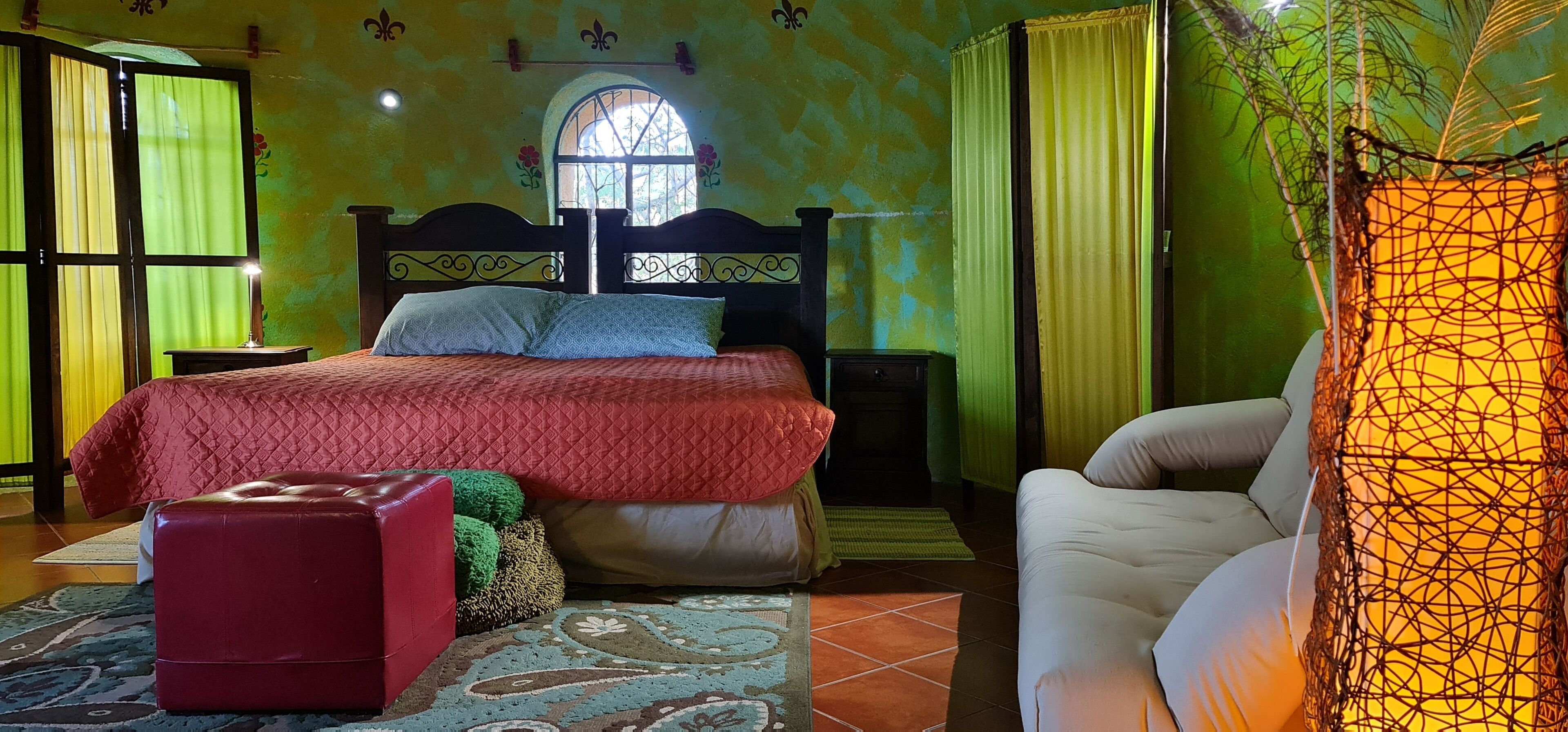 A bedroom with a bed, a red ottoman, a white chair, a nightstand, and a lamp.