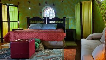 A bedroom with a bed, a red ottoman, a white chair, a nightstand, and a lamp.