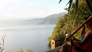 A person sitting on a wooden platform overlooking a lake with a stone structure nearby. - Atitlan Oasis Eco Hotel and Spa Kaalpul (San Antonio Palopo)