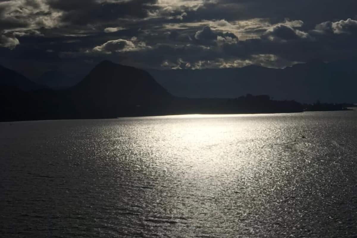 Ruhige Wasseroberfläche spiegelt das Sonnenlicht wider, im Hintergrund Berge und ein bewölkter Himmel.