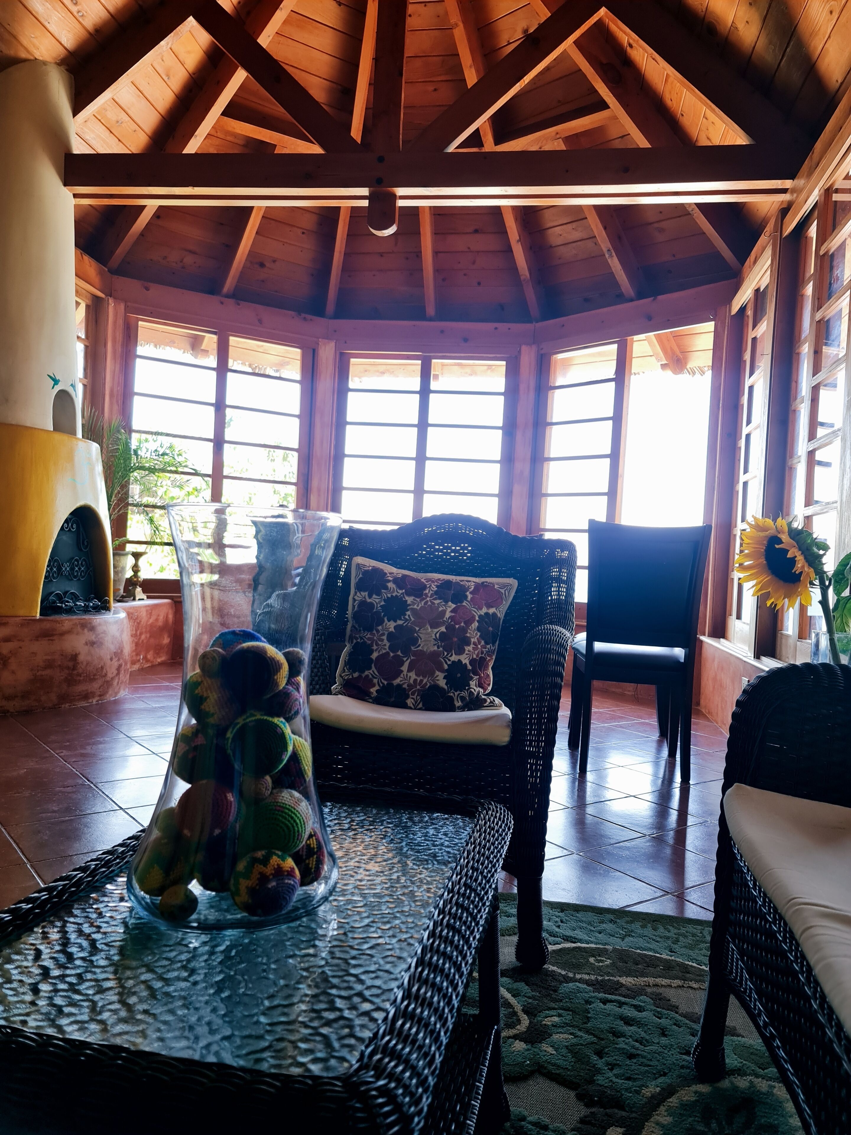 A cozy room with a high ceiling, wooden beams, a fireplace, wicker furniture, and large windows.