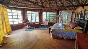 A rustic room with a bed, a chair, and a table. There are large windows with a view of greenery outside. - Atitlan Oasis Eco Hotel and Spa Kaalpul (San Antonio Palopo)