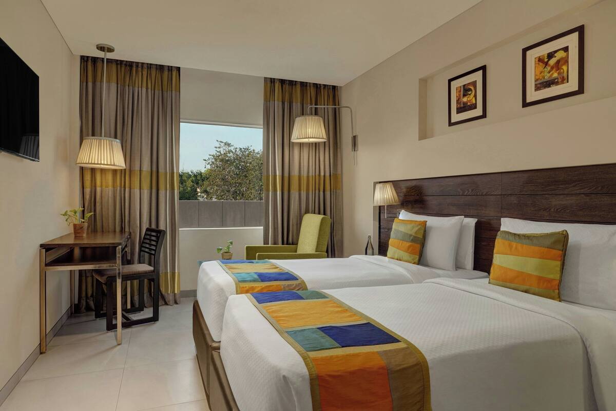 A hotel room with two beds, a desk, a chair, a TV, and a window with curtains.