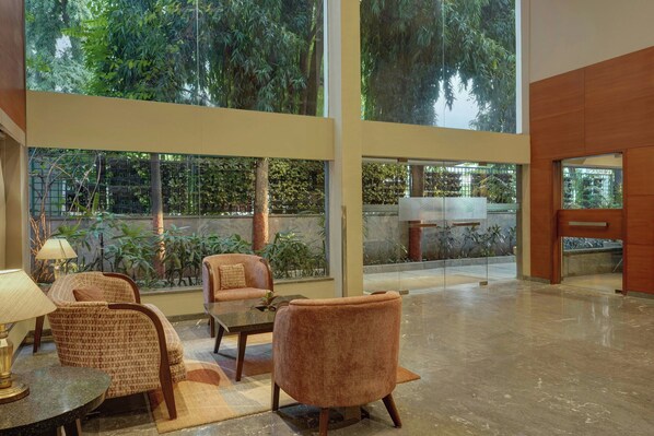A spacious lobby with high ceilings, large windows, and a view of lush greenery. There are comfortable seating areas with sofas and chairs.