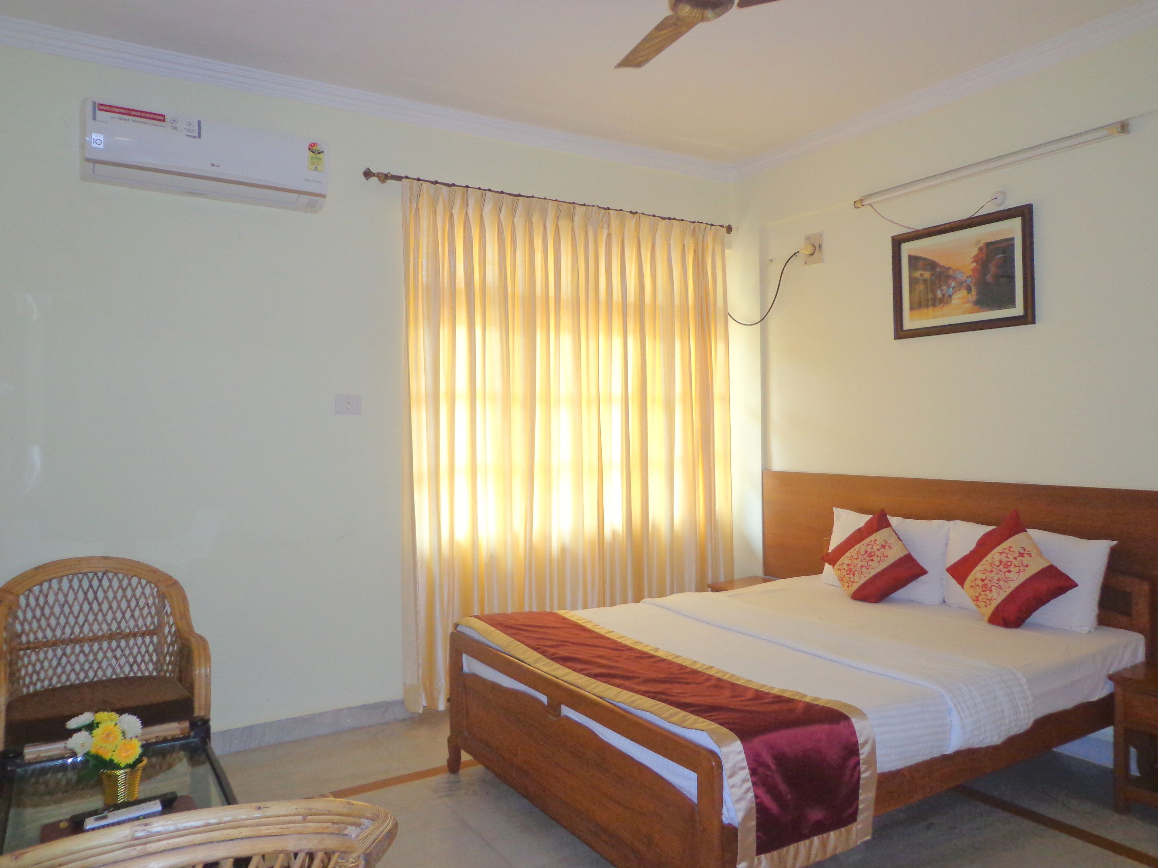 A bedroom with a wooden bed, a ceiling fan, an air conditioner, a chair, a table with a vase of flowers, and a window with curtains.