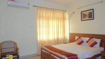 Une chambre avec un lit en bois, un ventilateur de plafond, un climatiseur, une chaise, une table ornée d’un vase de fleurs et une fenêtre avec des rideaux.