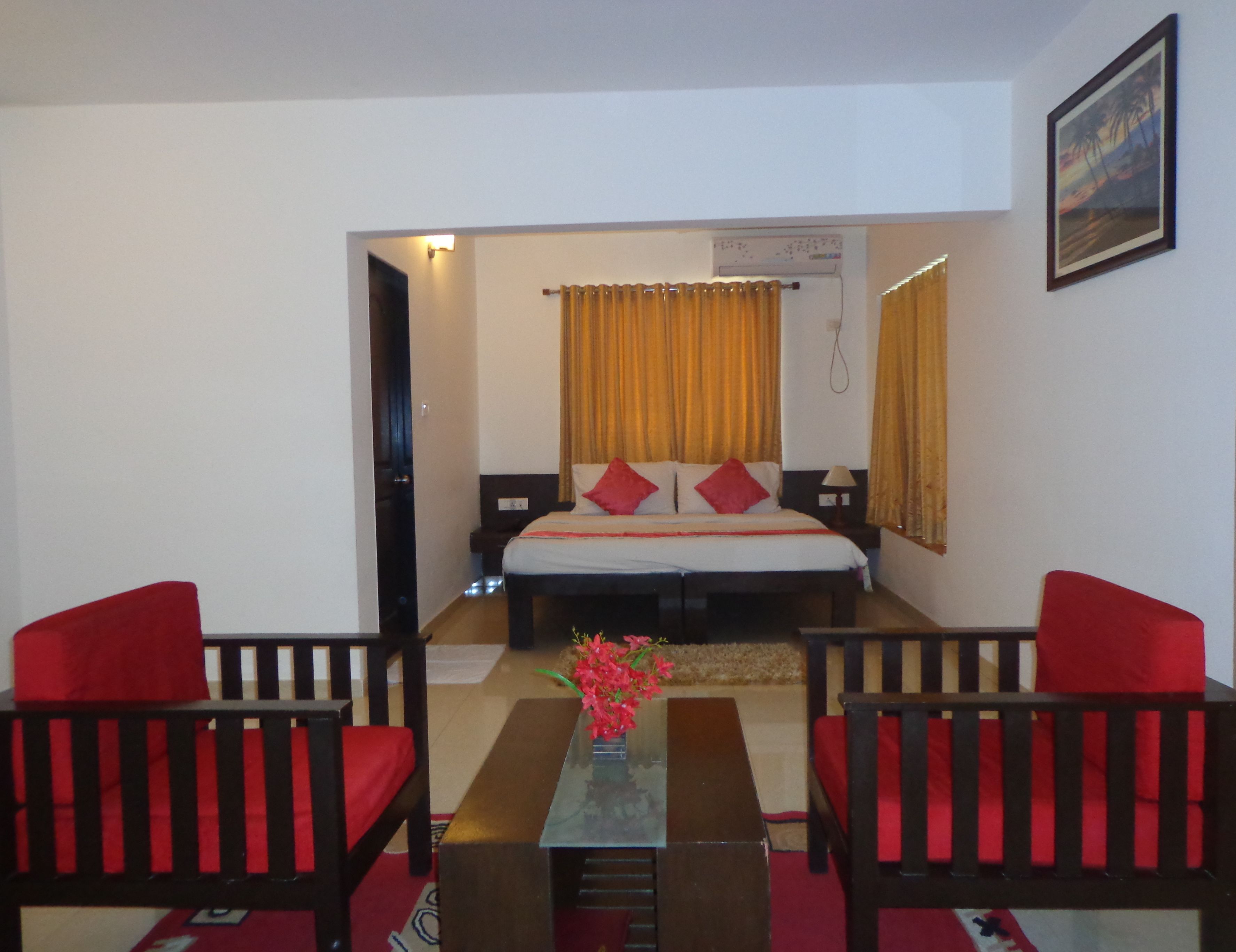 A hotel room with a bed, two red upholstered chairs, a small table with a vase of flowers, and a painting on the wall.