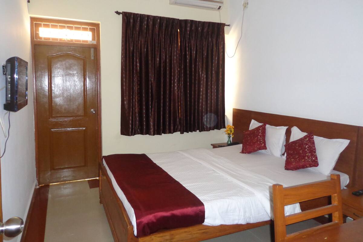 A room with a wooden bed, a red throw blanket, a white pillow, a dark curtain, and a wall-mounted air conditioner.