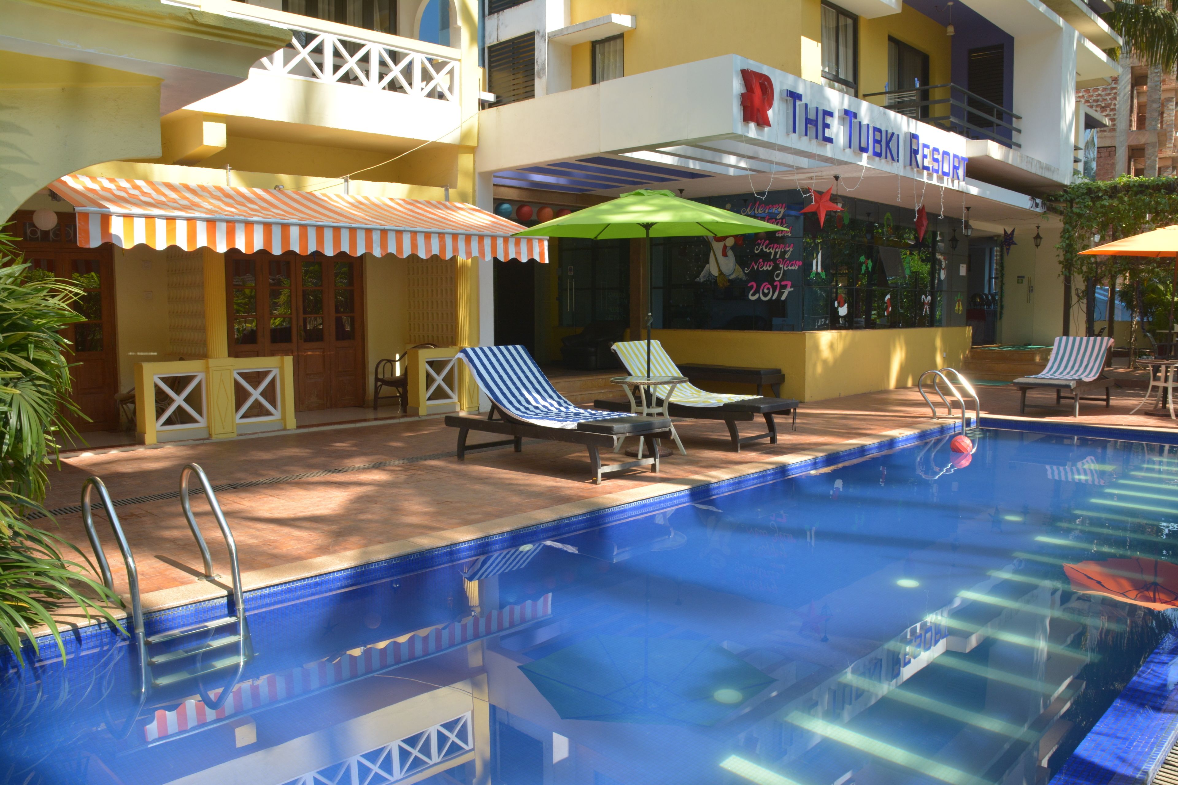 Una piscina con sillas de descanso y sombrillas, un bar junto a la piscina y un edificio con un letrero que dice "The Tubki Resort".