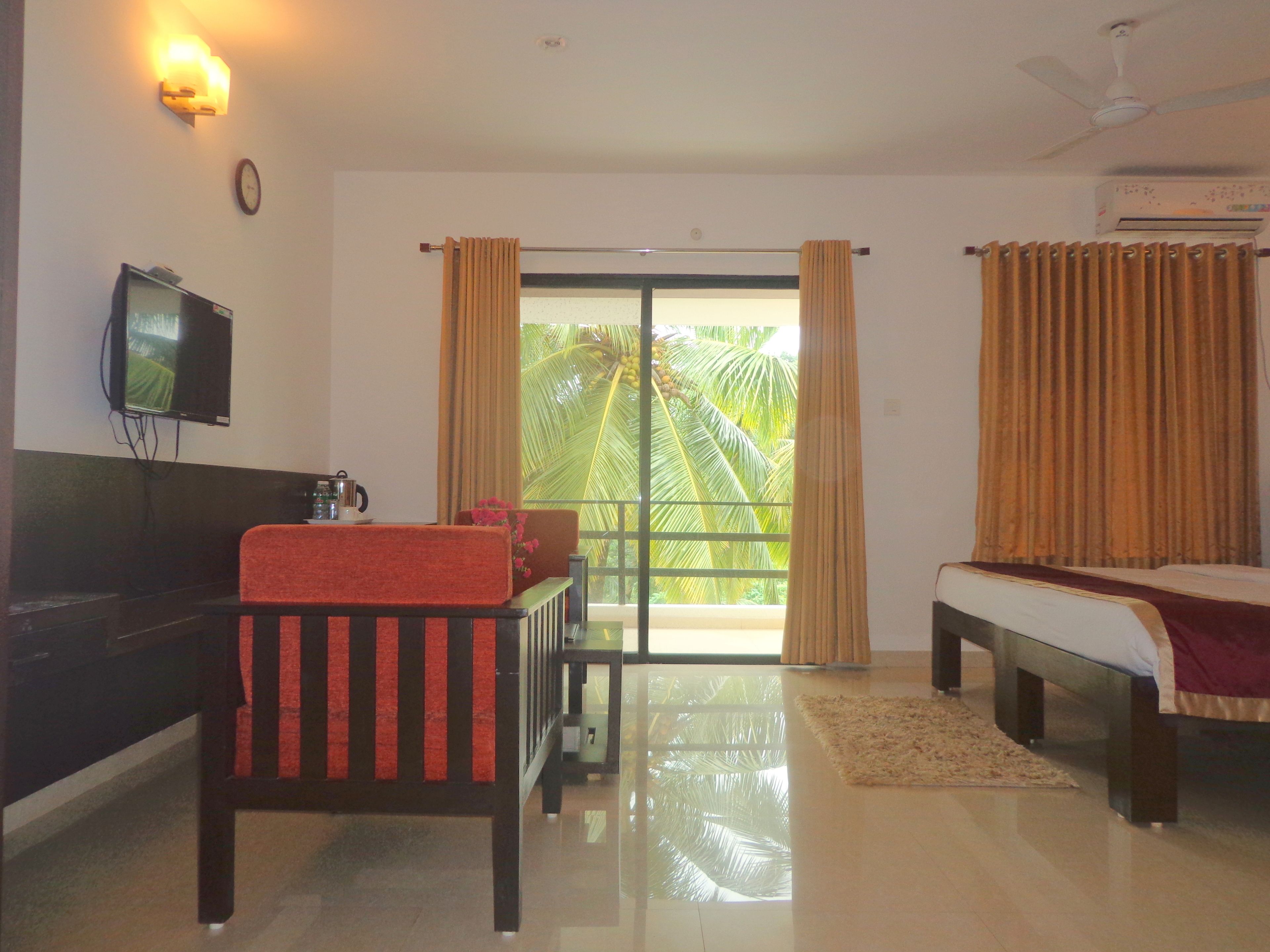 A hotel room with a bed, a sofa, a TV, a balcony with curtains, and a ceiling fan.