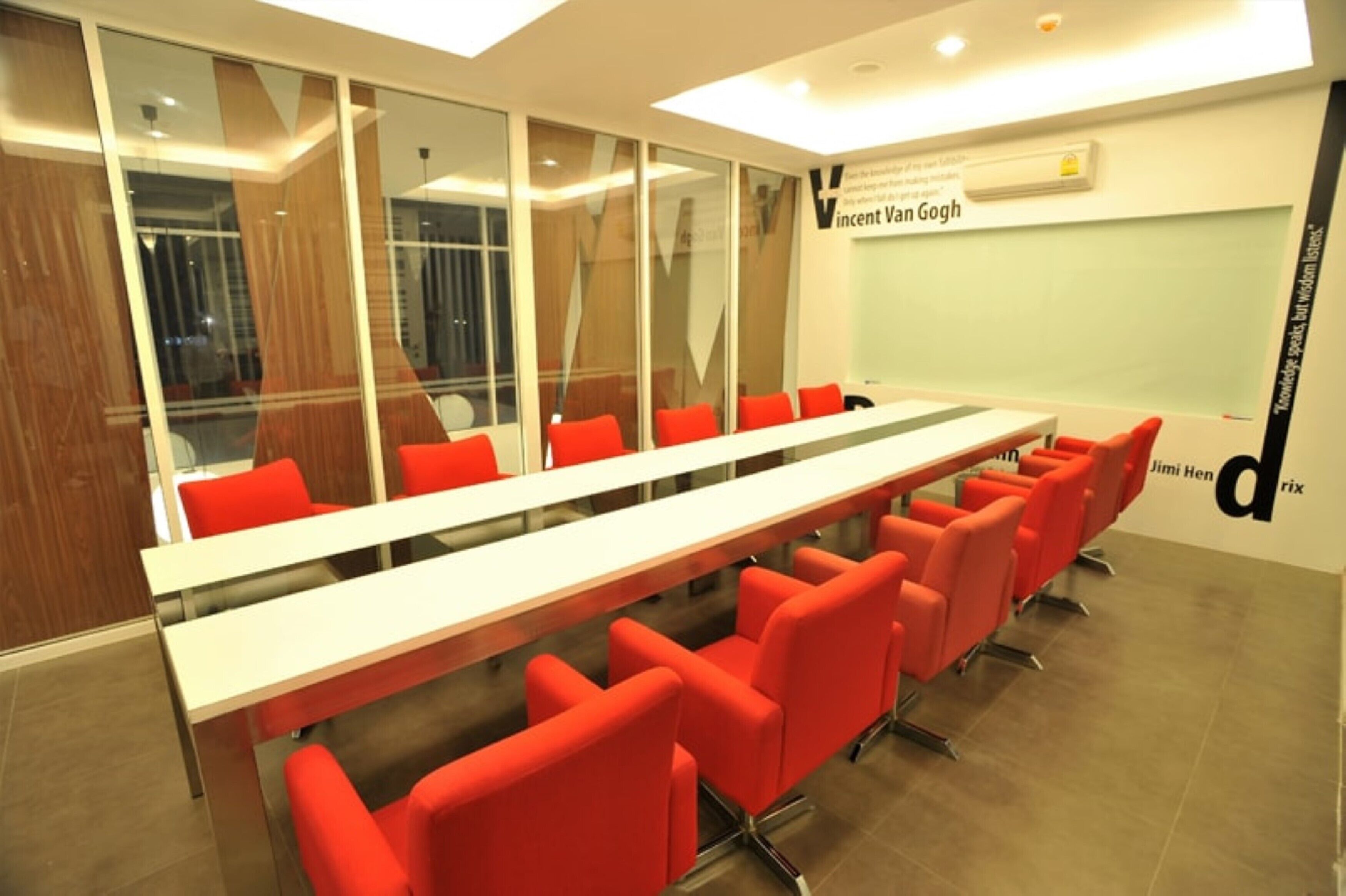 A conference room with a long table and red chairs, large windows, and a whiteboard with text.