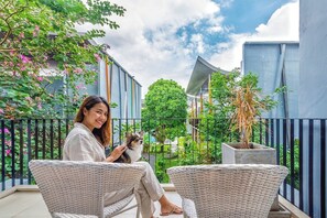 A balcony with wicker furniture, a potted plant, and a view of a garden and buildings. - Paeva Luxury Serviced Residence SHA (Bang Sao Thong)