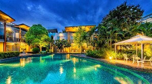 Una piscina con borde de baldosas, rodeada de exuberante vegetación y edificios modernos.