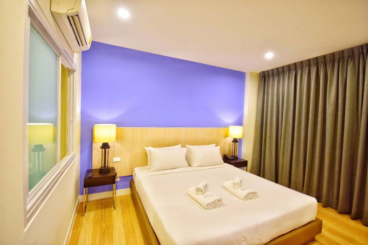 A hotel room with a bed, two bedside lamps, a window with curtains, and an air conditioning unit.
