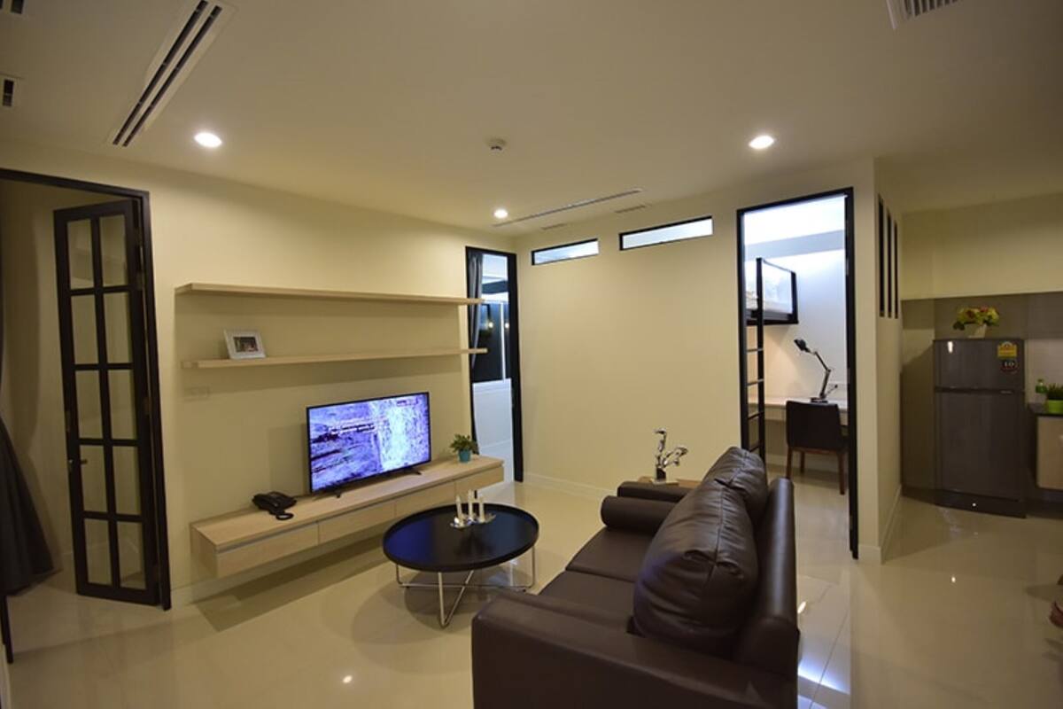 A modern living room with a flat-screen TV, a sofa, a coffee table, and a kitchen area visible through an open door.