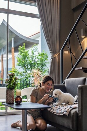 A person sitting on a sofa with a small dog, a table with a candle and a vase, and a view of greenery outside. - Paeva Luxury Serviced Residence SHA (Bang Sao Thong)