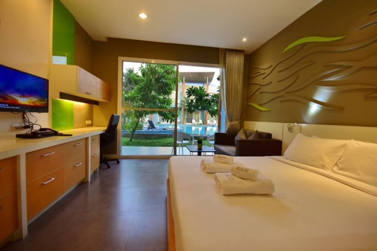 A hotel room with a bed, a desk, a TV, and a view of a pool and greenery.