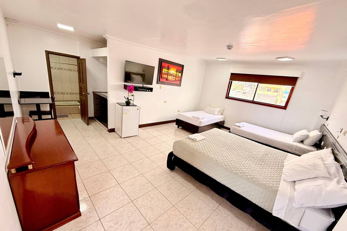 Habitación de hotel con cama, escritorio, refrigerador pequeño, televisión y ventana con vista a edificios.