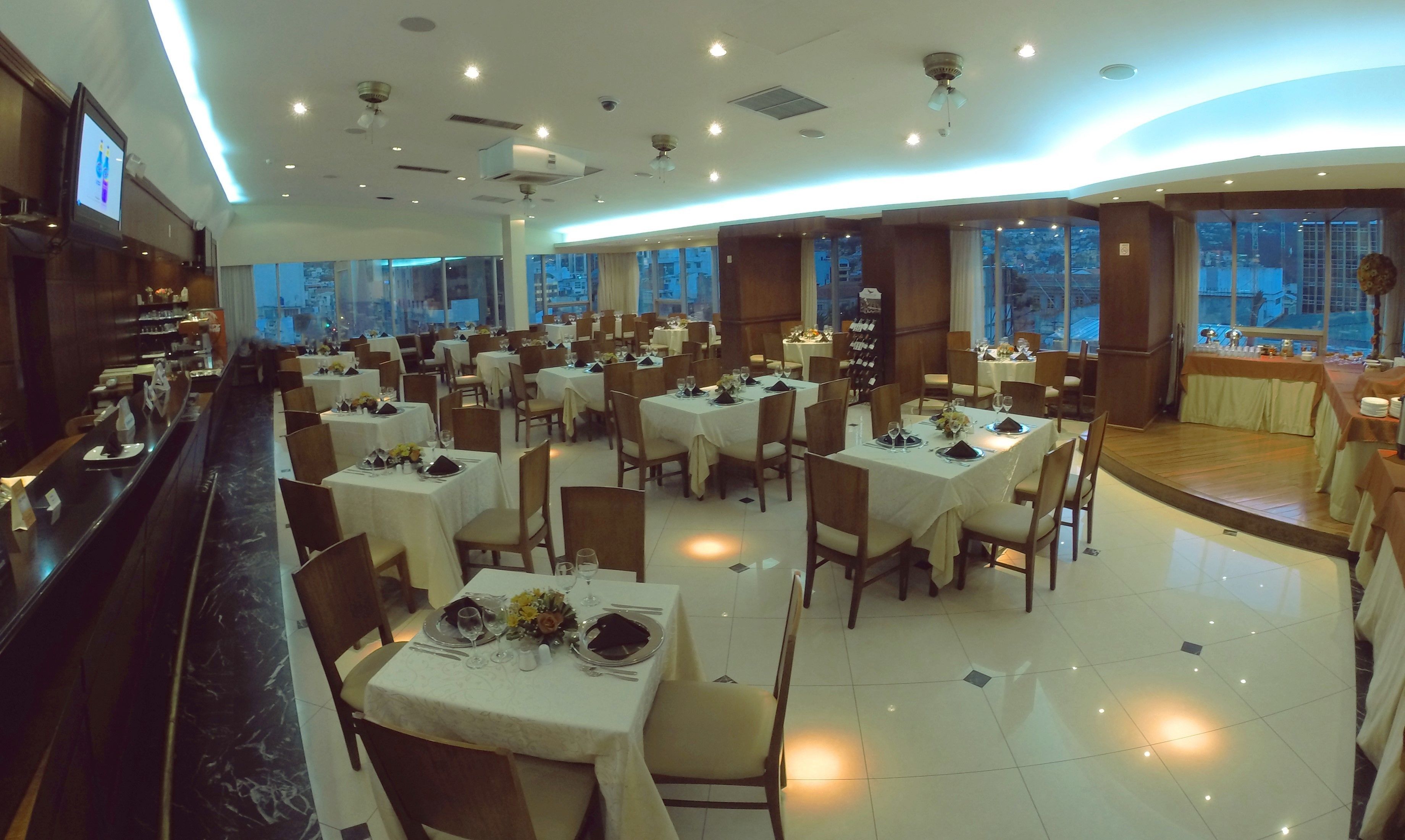 A spacious restaurant with multiple tables set for dining, large windows offering city views, and a modern ceiling design with recessed lighting.