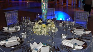 A round table set for a formal event, with a tall vase of flowers and a blue-lit backdrop. - Hotel Emperador (Ambato)