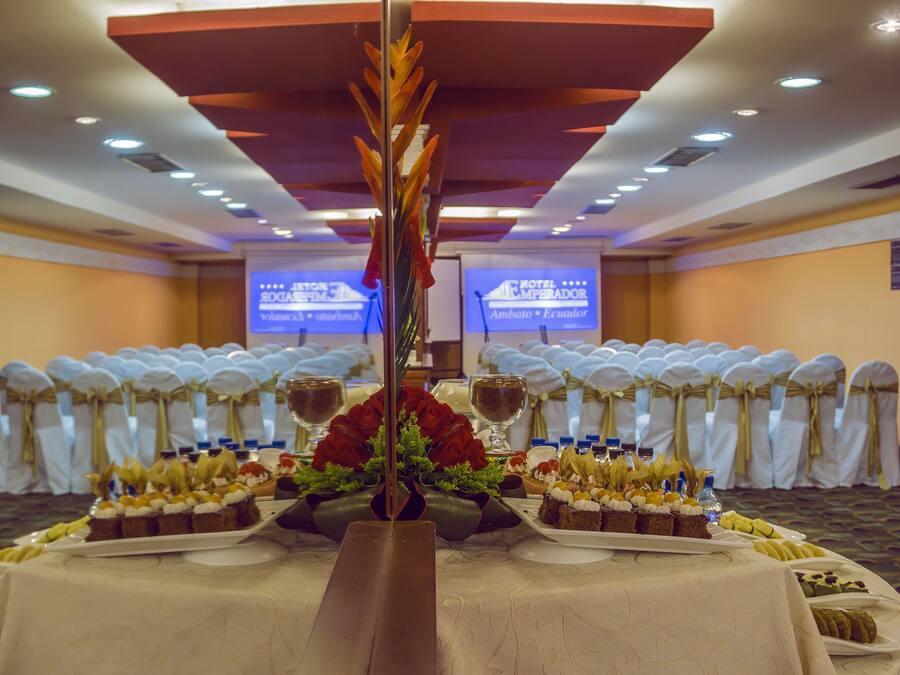 Uma sala de banquetes com fileiras de cadeiras brancas, uma mesa central com um arranjo floral e uma tela de projeção ao fundo.