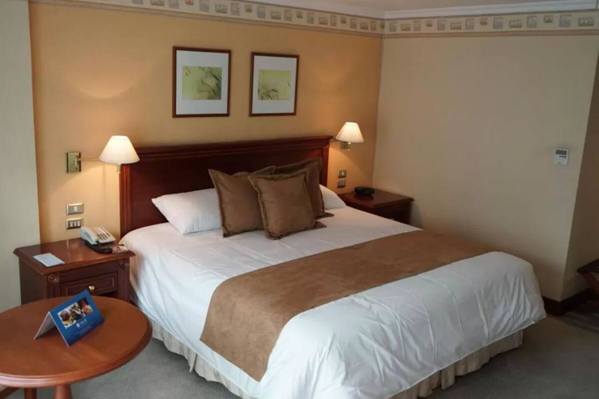 A hotel room with a large bed, two bedside lamps, a wooden nightstand with a phone, a small round table with a brochure, and two framed pictures on the wall.