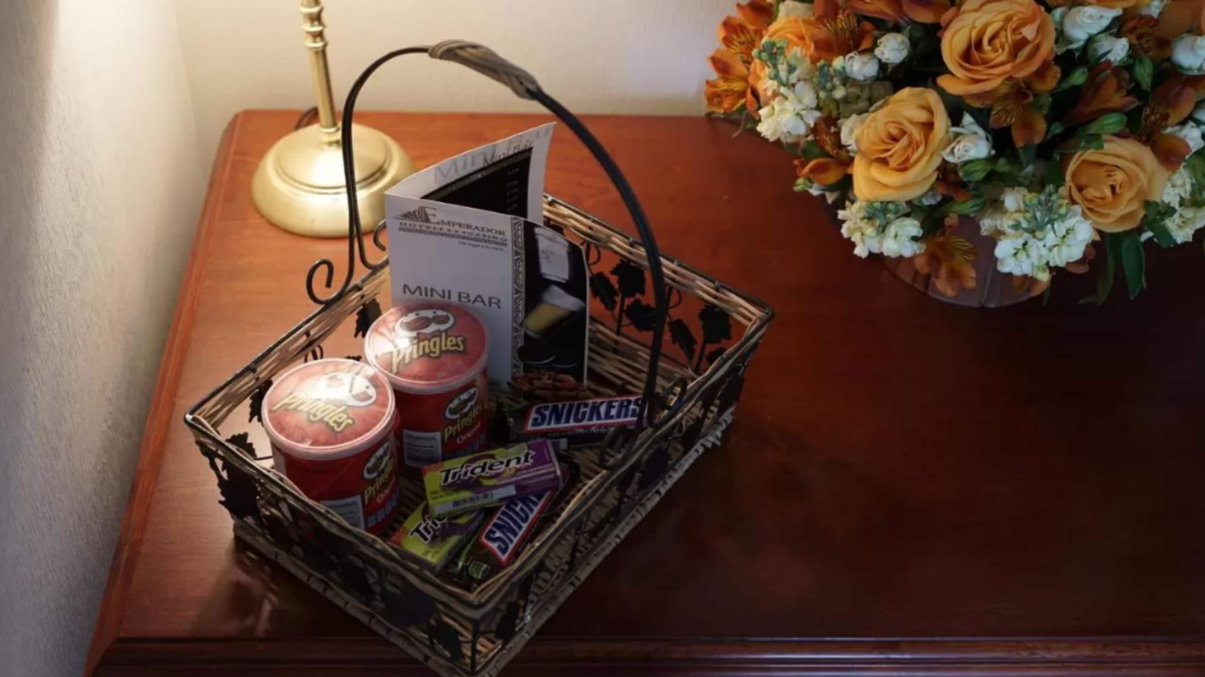 A wooden table with a basket containing snacks, a mini bar sign, and a bouquet of flowers.