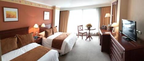 Quarto de hotel com duas camas, uma escrivaninha com televisão e uma janela com cortinas.