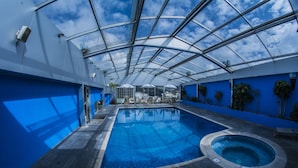 A large indoor swimming pool with a glass ceiling, surrounded by blue walls and potted plants. - Hotel Emperador (Ambato)