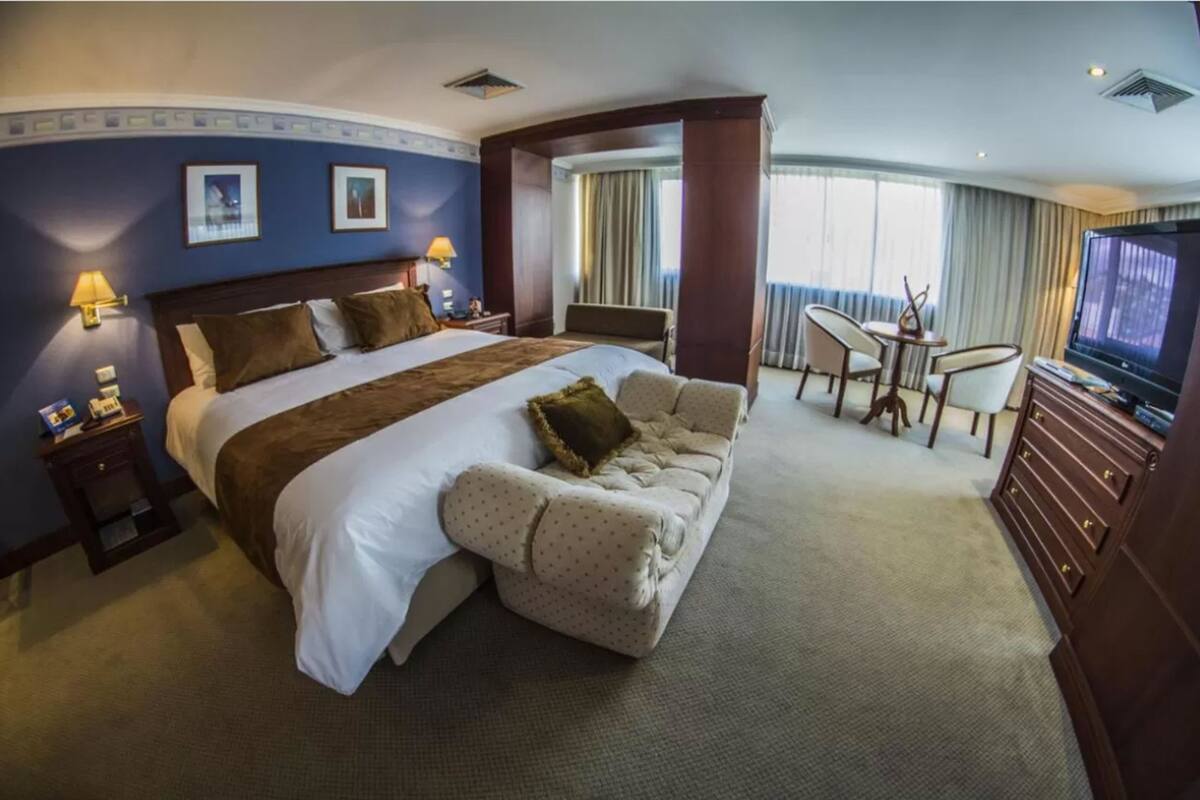 A hotel room with a large bed, a sofa, a TV, and a dining area.