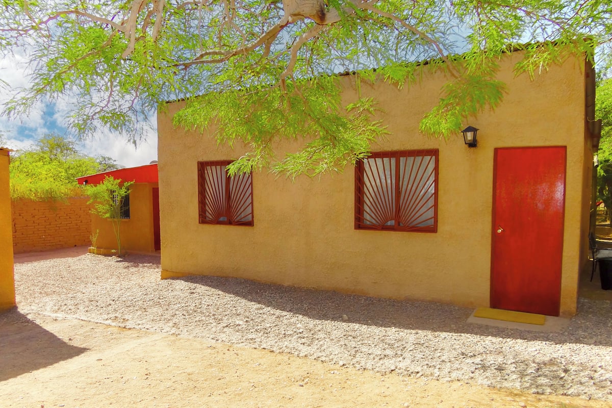 A building with two windows and a red door, surrounded by gravel and greenery.