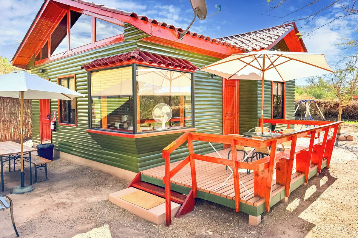 A small, green wooden cabin with a red roof, a satellite dish, and a wooden deck with outdoor furniture.