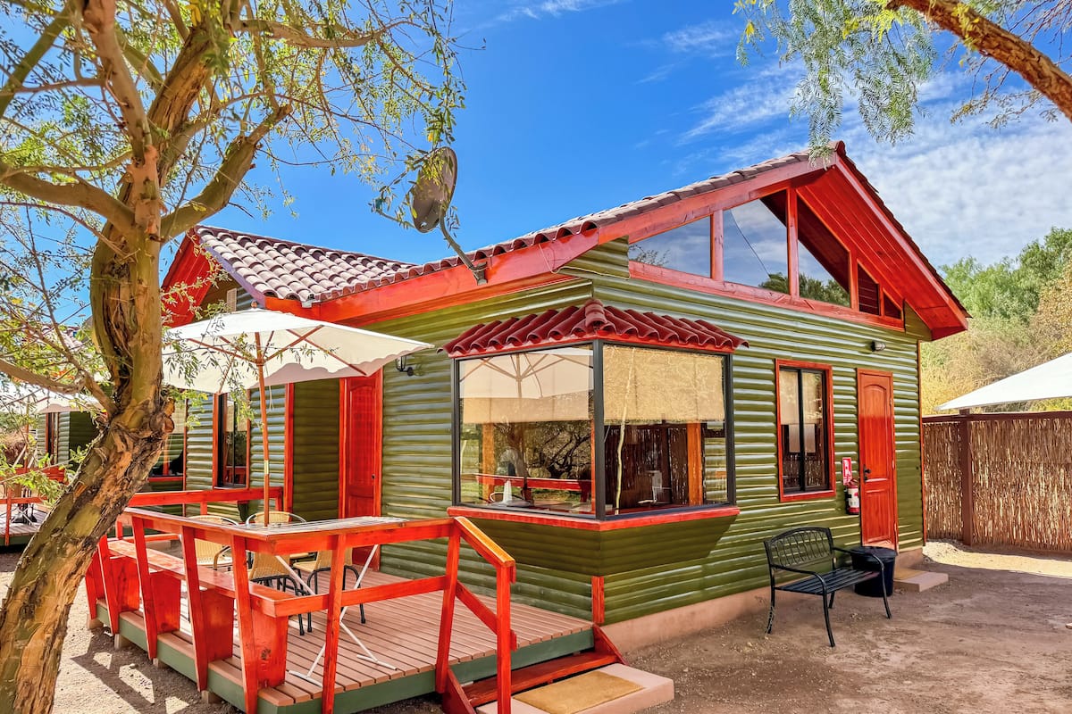 A rustic cabin with a wooden deck, red accents, and a large window.