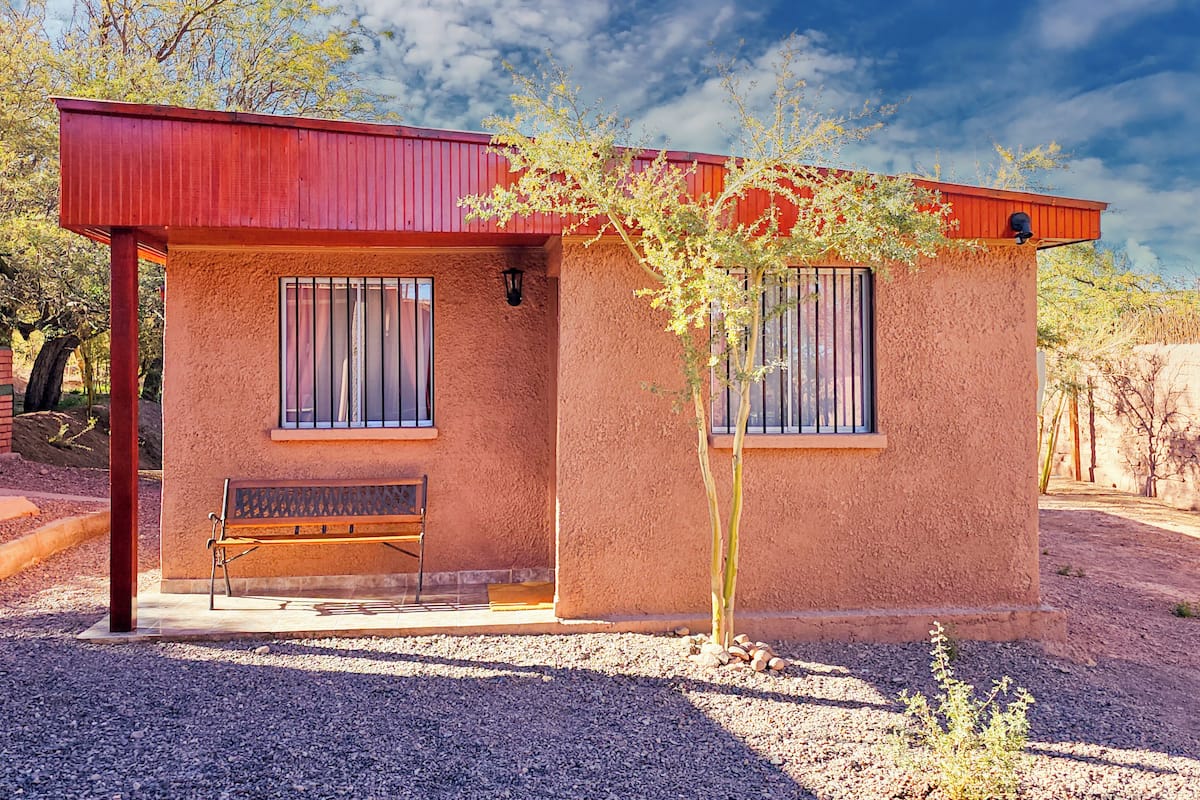 A small, single-story building with a red roof and beige walls, featuring two windows and a bench outside.