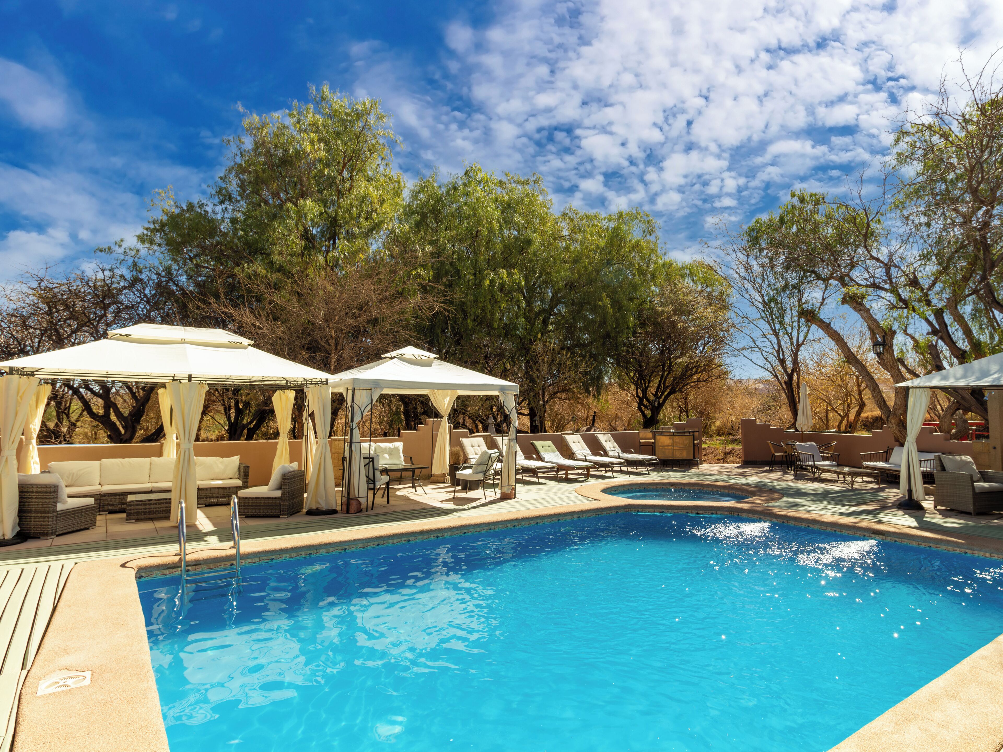 Un área de piscina con sillones de descanso y sombrillas, rodeada de árboles.