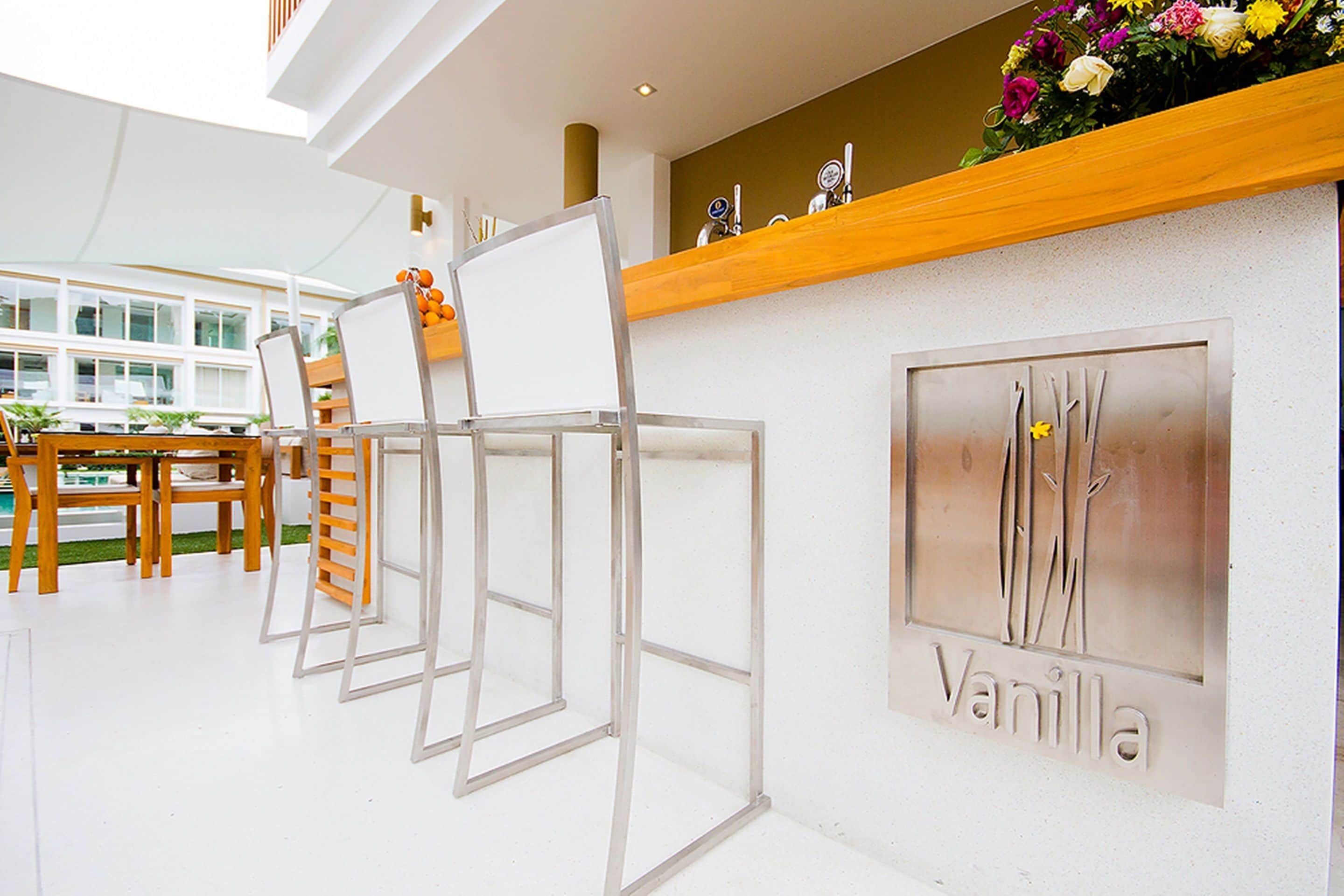 Un interior moderno con una zona de bar que muestra un letrero 'Vanilla', sillas blancas y una barra de madera con objetos decorativos.