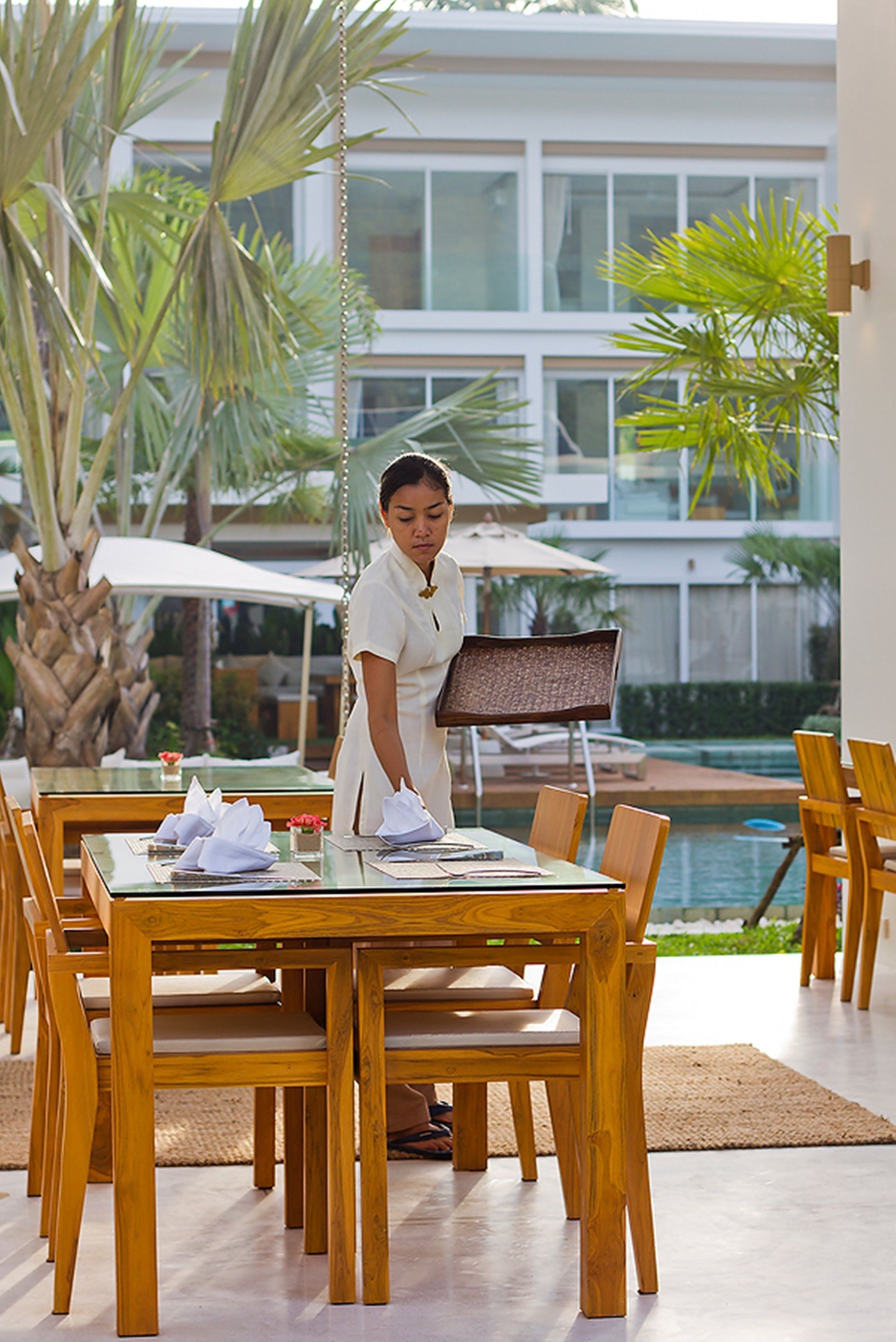 A hotel dining area with wooden tables and chairs, a pool in the background, and a palm tree.