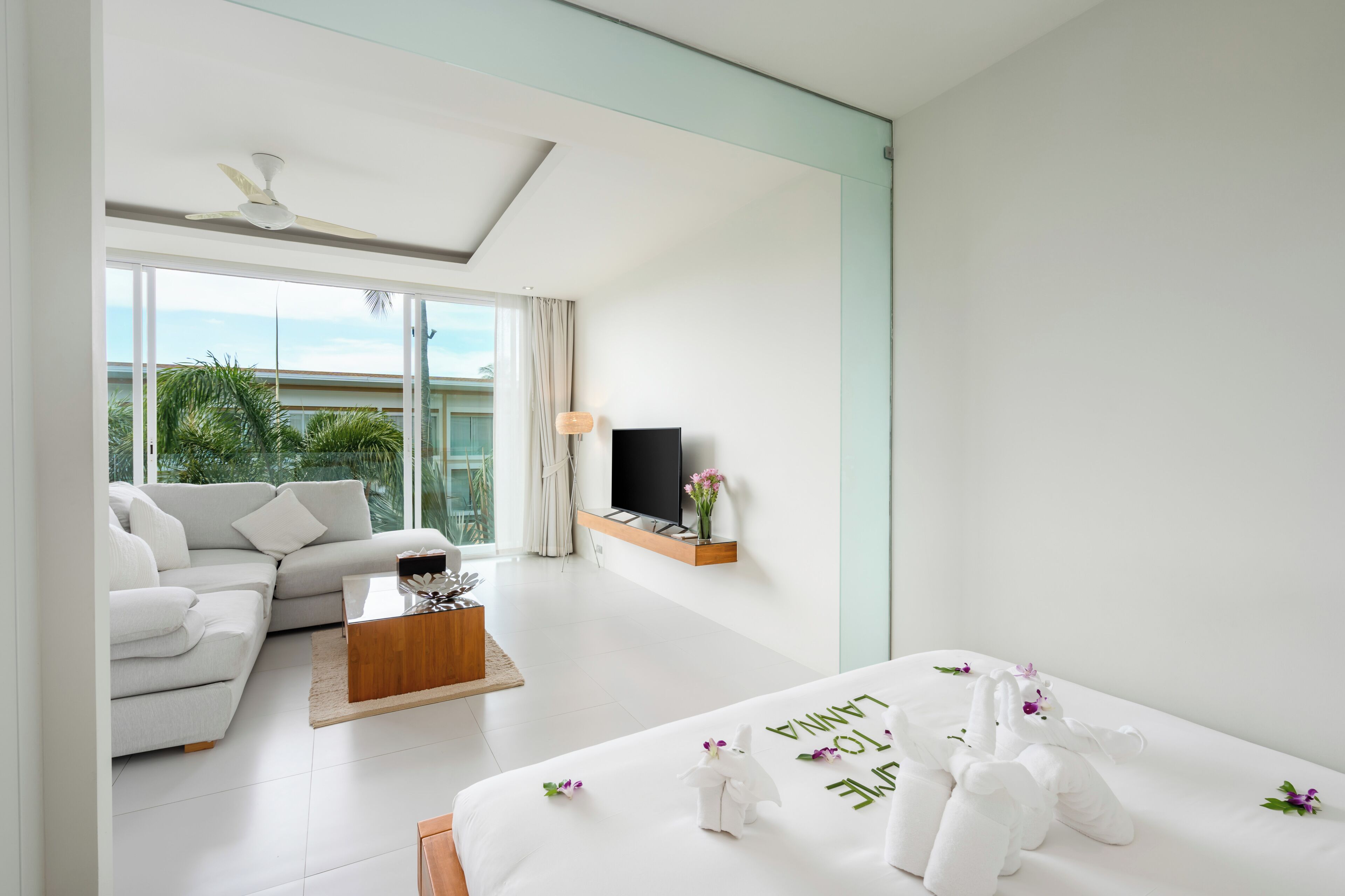 Habitación de hotel moderna con sofá, mesa de centro, cama con toalla y arreglo floral, televisor y balcón con vistas a follaje.
