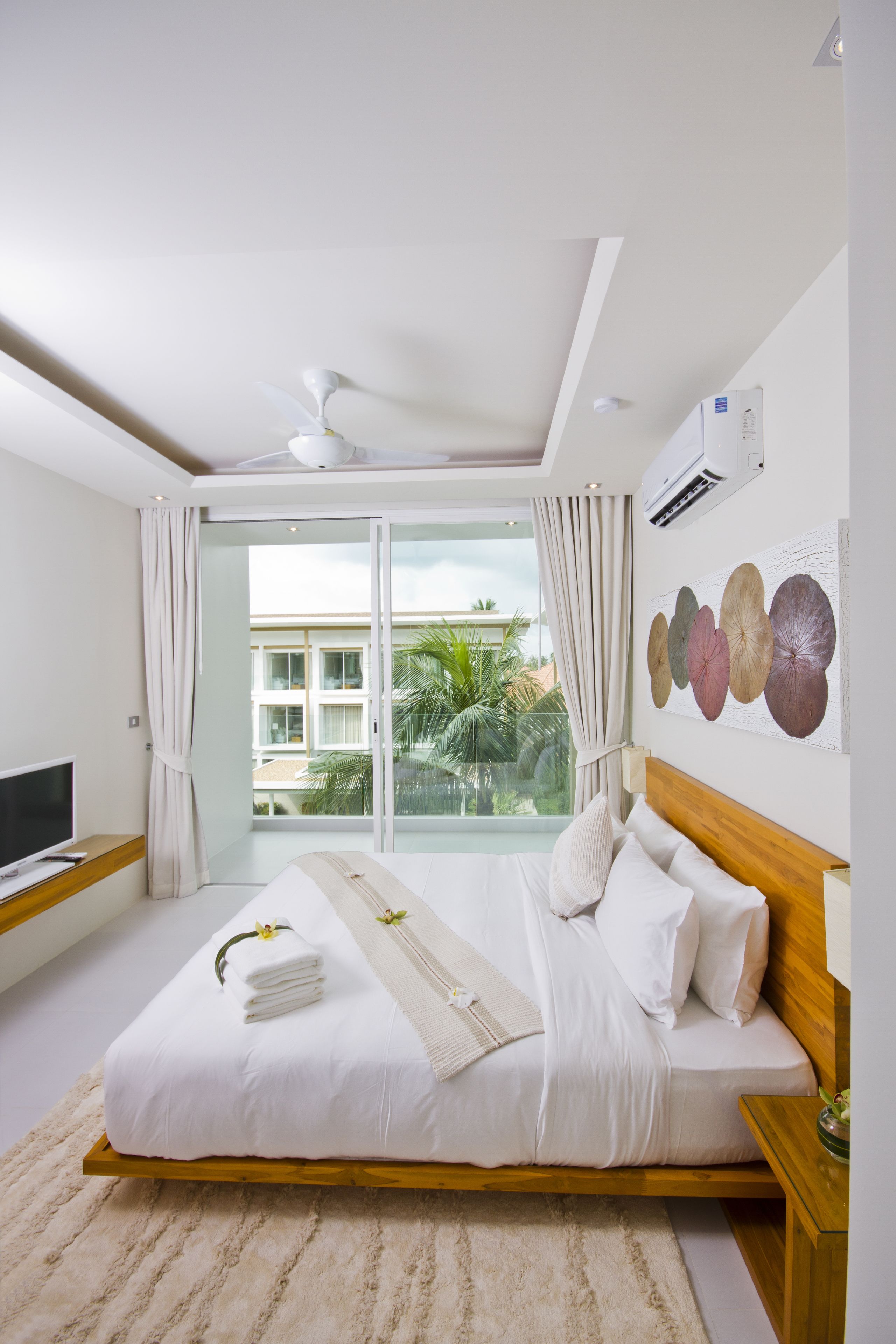 A modern hotel room with a large bed, a flat-screen TV, a ceiling fan, and a view of palm trees and buildings through the glass door.