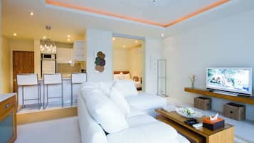 A modern living room with a white sofa, a wooden coffee table, and a flat-screen TV. There is a kitchen area with white cabinets and a bar stool.