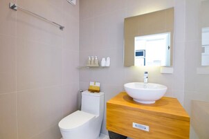 Un baño moderno con un inodoro blanco, un tocador de madera con lavamanos blanco y un espejo sobre el lavamanos. Hay una repisa con artículos de aseo y un perchero en la pared.