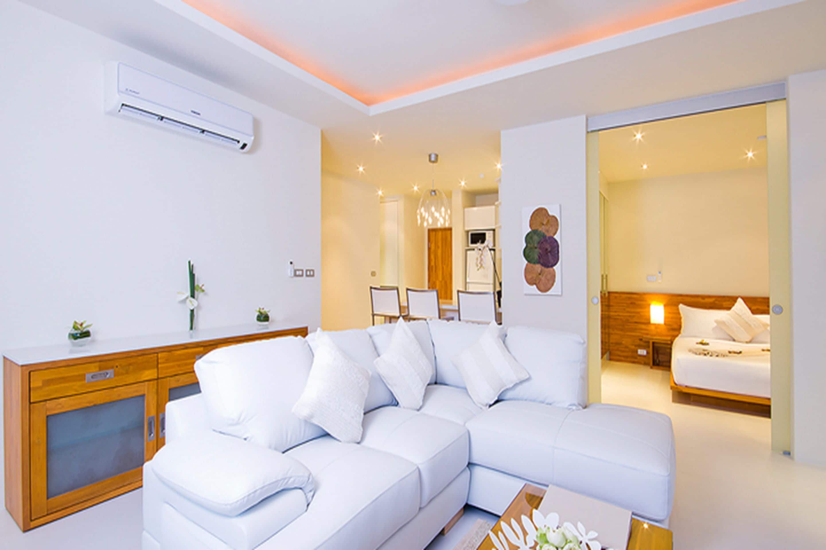A modern living room with a white sectional sofa, a wooden cabinet, and a bed with a wooden headboard.