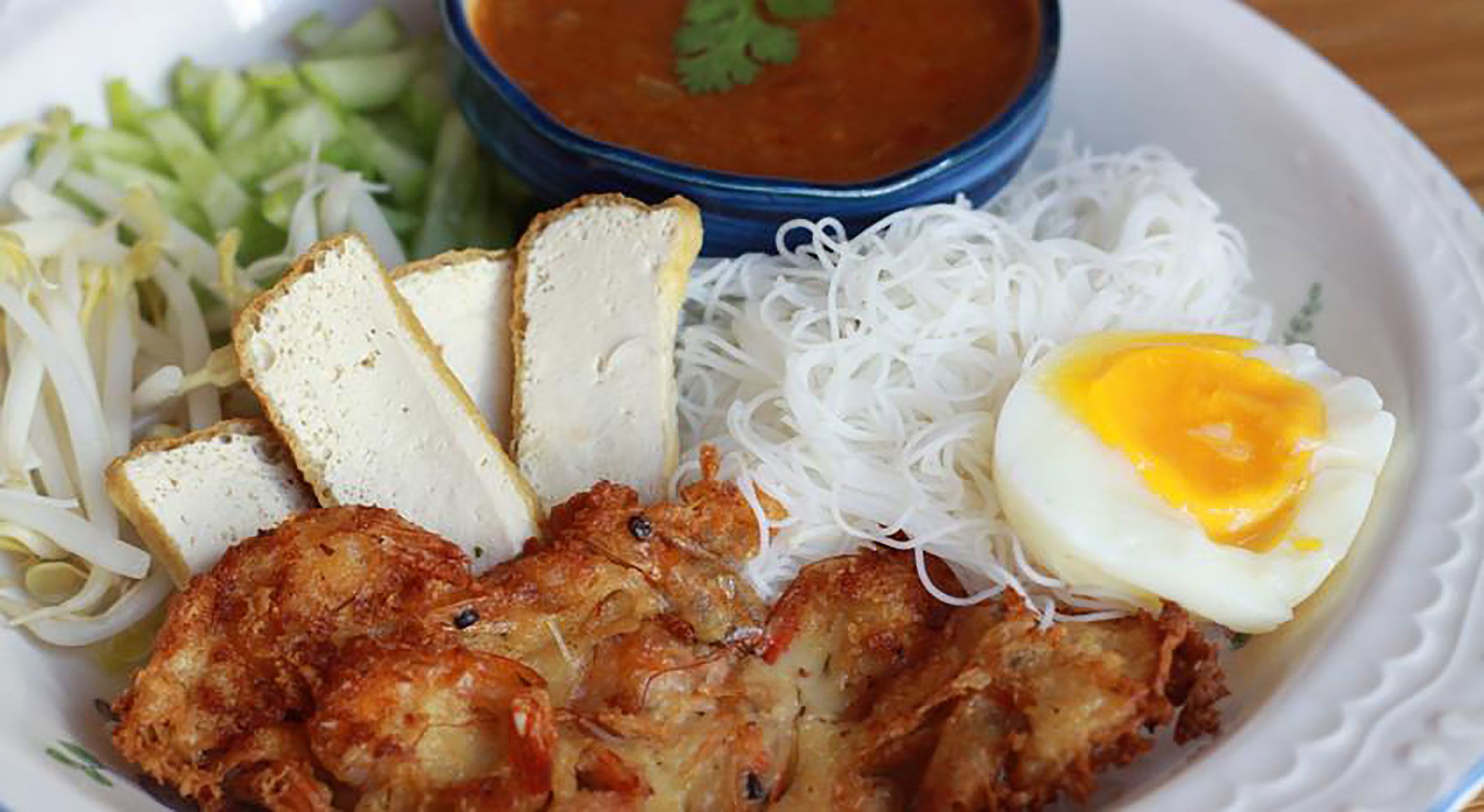 A plate with fried chicken, noodles, sliced bread, a boiled egg, and a bowl of sauce.