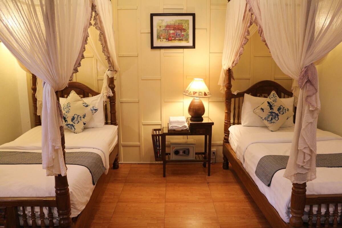 A room with two wooden beds, each with a canopy, a bedside table with a lamp, and a framed picture on the wall.